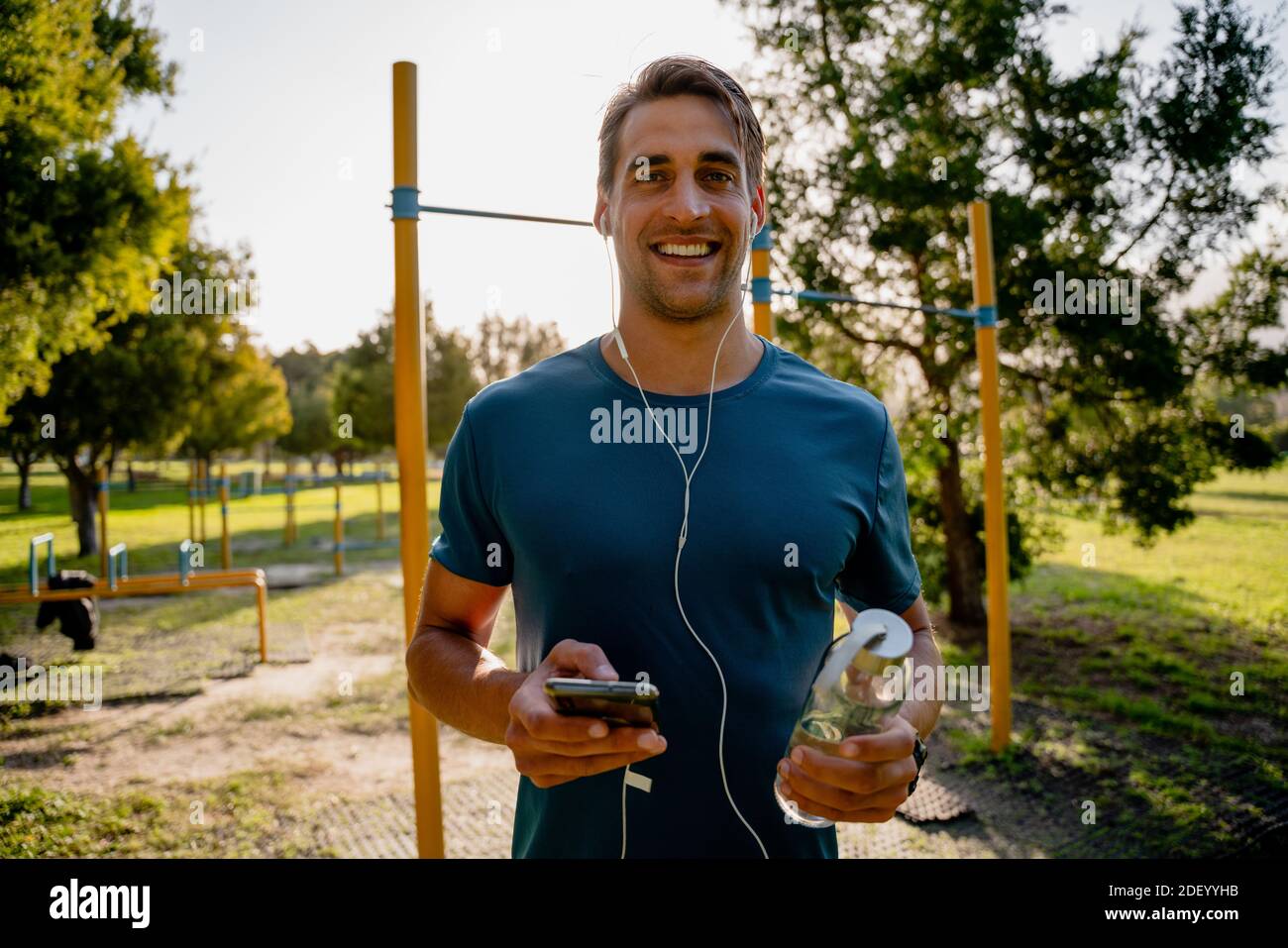 Beau jeune athlète masculin s'exerçant à l'extérieur en écoutant de la musique écouteurs avec smartphone et bouteille d'eau Banque D'Images
