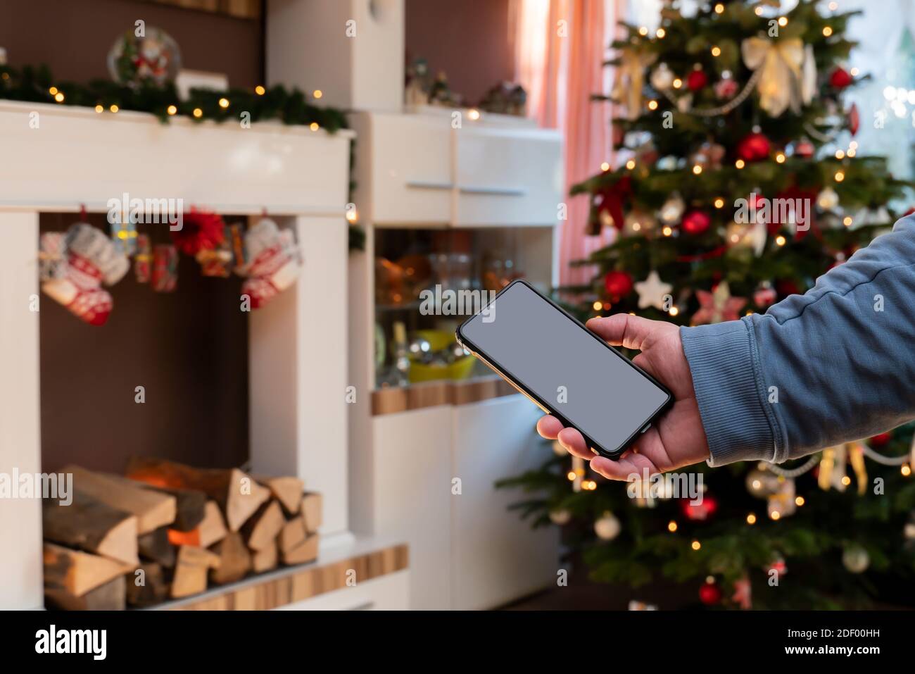 Une main mâle tient un smartphone. L'homme est debout dans un salon décoré de Christmassy. Console de l'arbre de Noël et de la cheminée. Banque D'Images