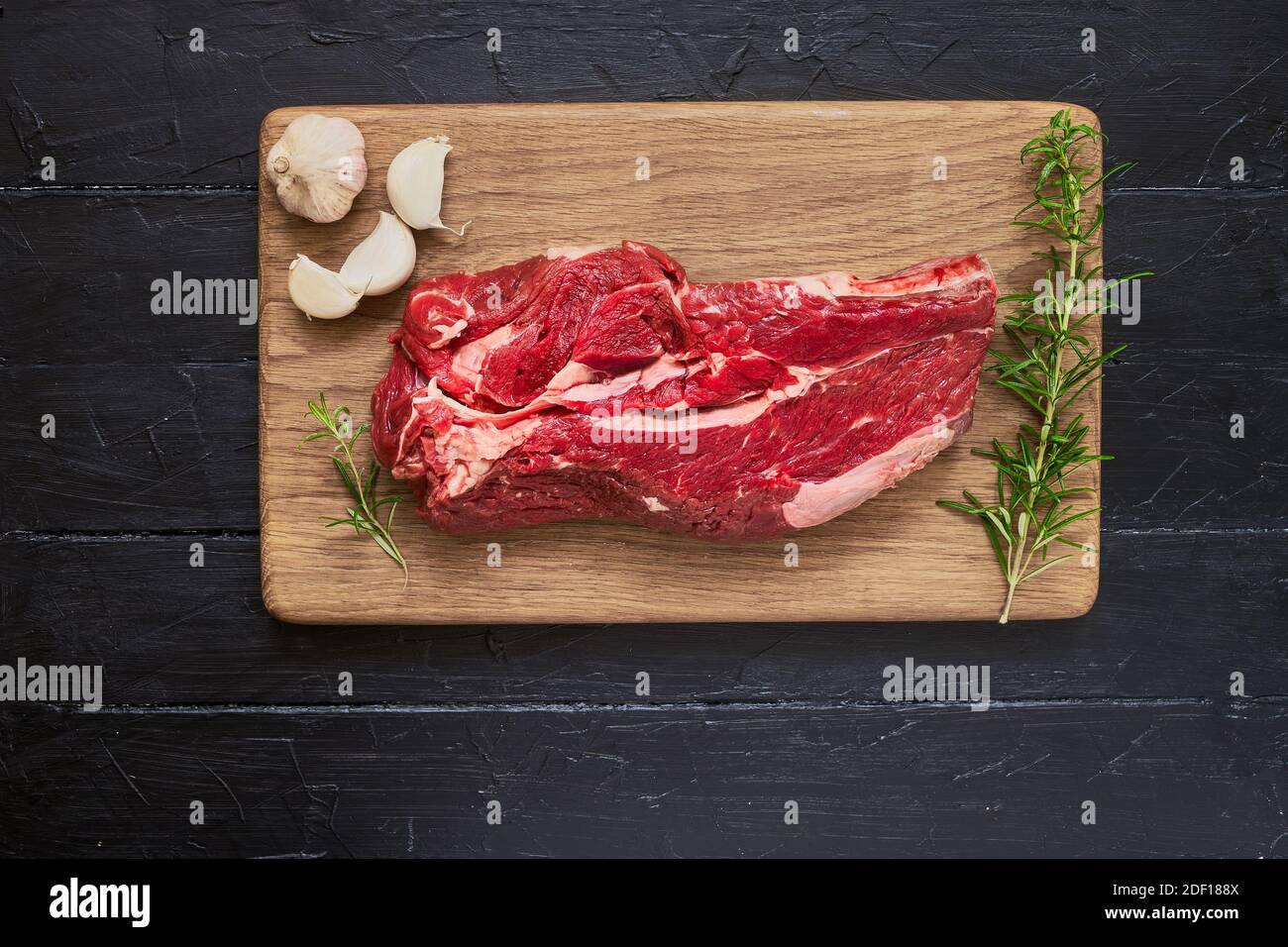 Viande de bœuf fraîche et crue à l'ail et au romarin sur la planche à découper sur une table en bois noir. Espace de copie, vue de dessus. Aliments biologiques. Banque D'Images