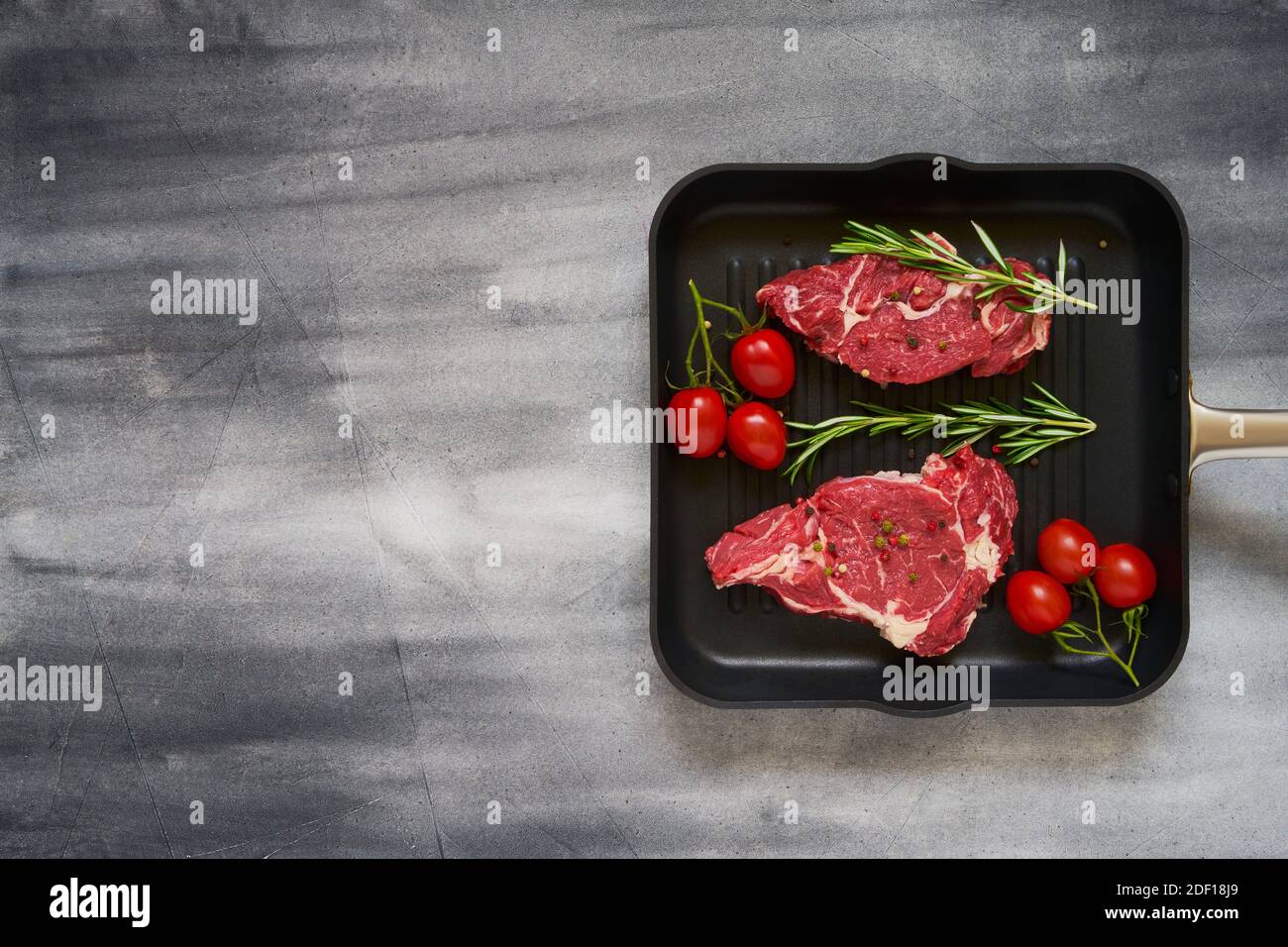 Steaks de bœuf frais crus au romarin, au poivre et aux tomates dans une casserole sur fond gris. Vue de dessus, espace de copie. Banque D'Images