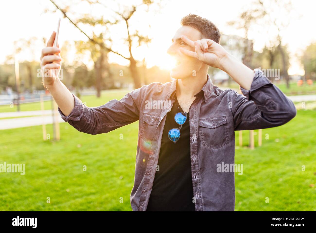 Portrait d'un homme en train de rire, portant des lunettes de soleil, debout dans un parc avec un smartphone. Il prend des photos de lui-même et montre le geste de paix sur c Banque D'Images