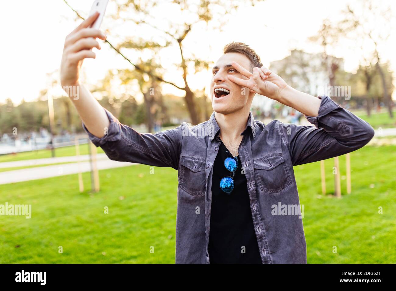 Portrait d'un homme en train de rire, portant des lunettes de soleil, debout dans un parc avec un smartphone. Il prend des photos de lui-même et montre le geste de paix sur c Banque D'Images