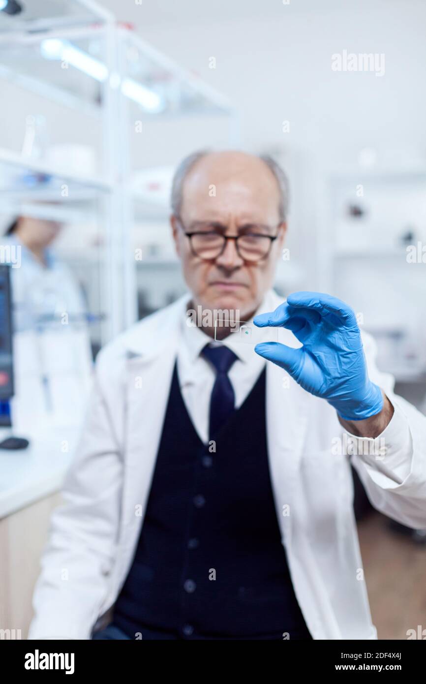 Un scientifique expérimenté dans un laboratoire très occupé tenant une lame de verre dans un laboratoire de microbiologie. Chercheur senior en laboratoire stérile regardant sur une lame de microscope portant une blouse de laboratoire. Banque D'Images