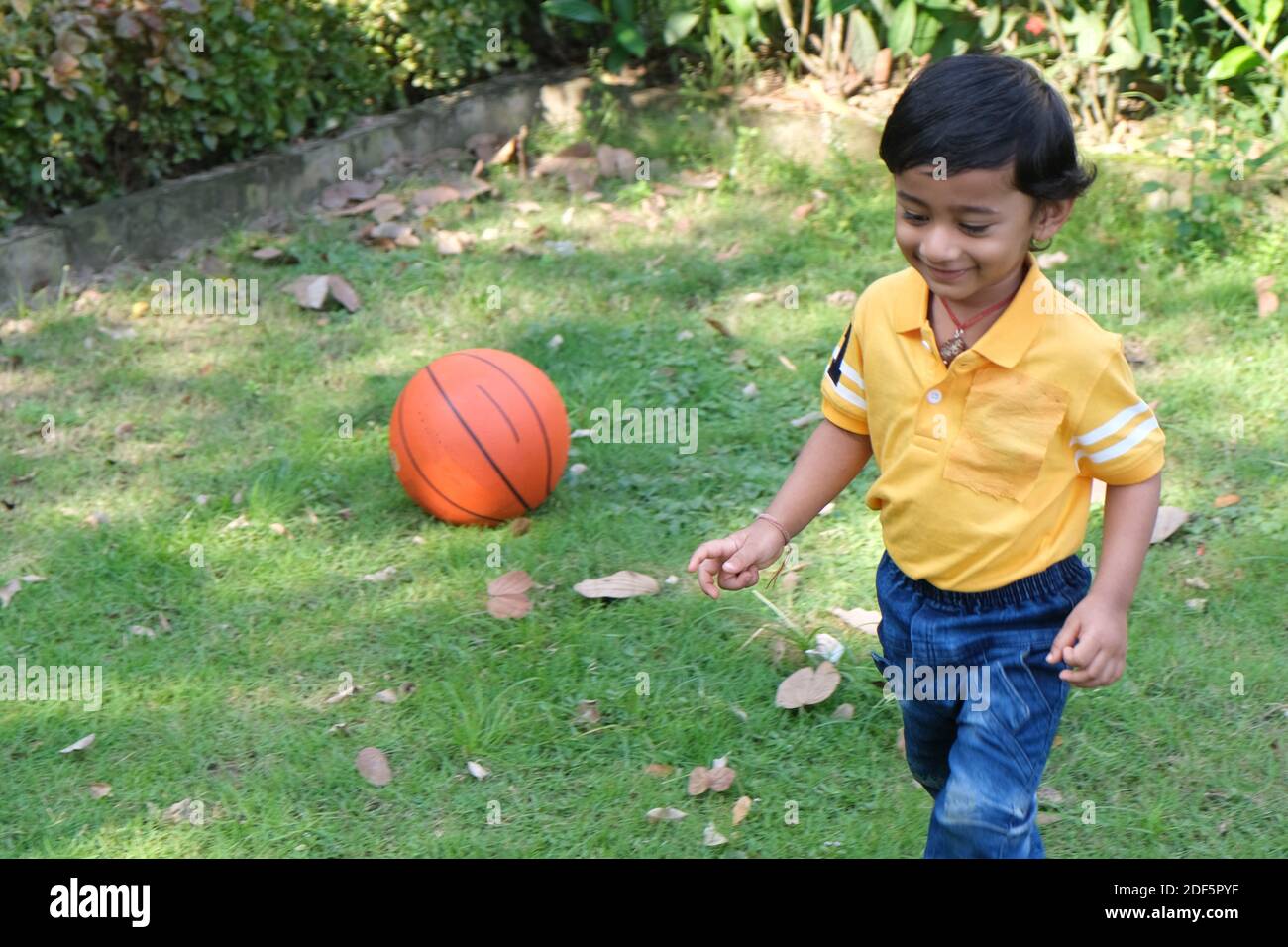 Petit garçon indien mignon gai brune tamoul dans vibrant jaune vêtements décontractés jouant avec lui-même avec une boule orange sur fond vert naturel Banque D'Images