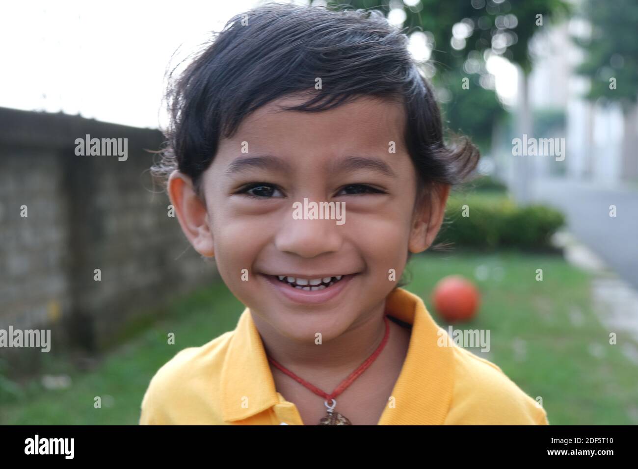 Petit garçon indien mignon gai brune tamoul dans vibrant jaune vêtements décontractés jouant avec lui-même avec une boule orange sur fond vert naturel Banque D'Images