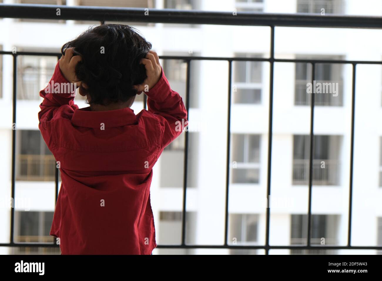 Portrait d'Indien mignon petit gai brune petit garçon tamoul porter une chemise rouge vif tout en étant debout sur un balcon à l'intérieur un fond urbain blanc Banque D'Images