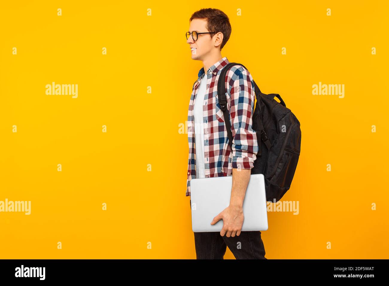 Un homme heureux avec des lunettes, un homme dans une chemise à carreaux, va avec un ordinateur portable fermé dans ses mains, sur un fond jaune Banque D'Images