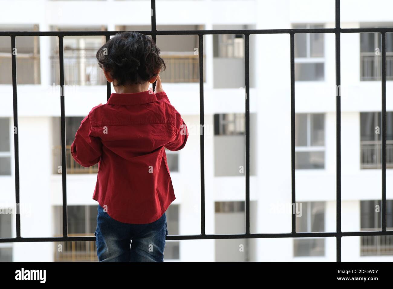 Portrait d'Indien mignon petit gai brune petit garçon tamoul porter une chemise rouge vif tout en étant debout sur un balcon à l'intérieur un fond urbain blanc Banque D'Images