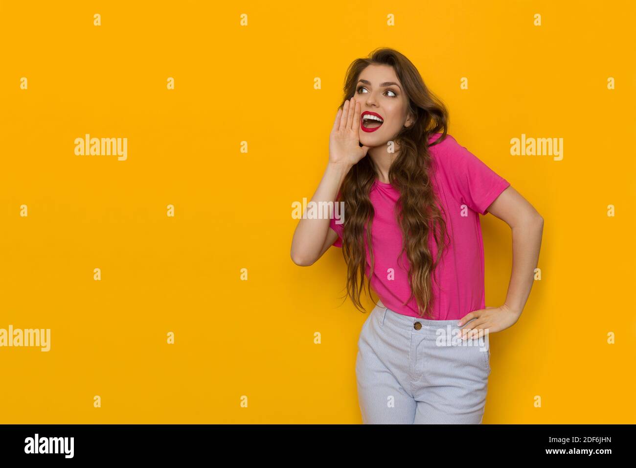 Une jeune femme heureuse en chemise rose tient la main sur le menton, regarde loin et crie. Trois quarts de long studio tourné sur fond jaune. Banque D'Images