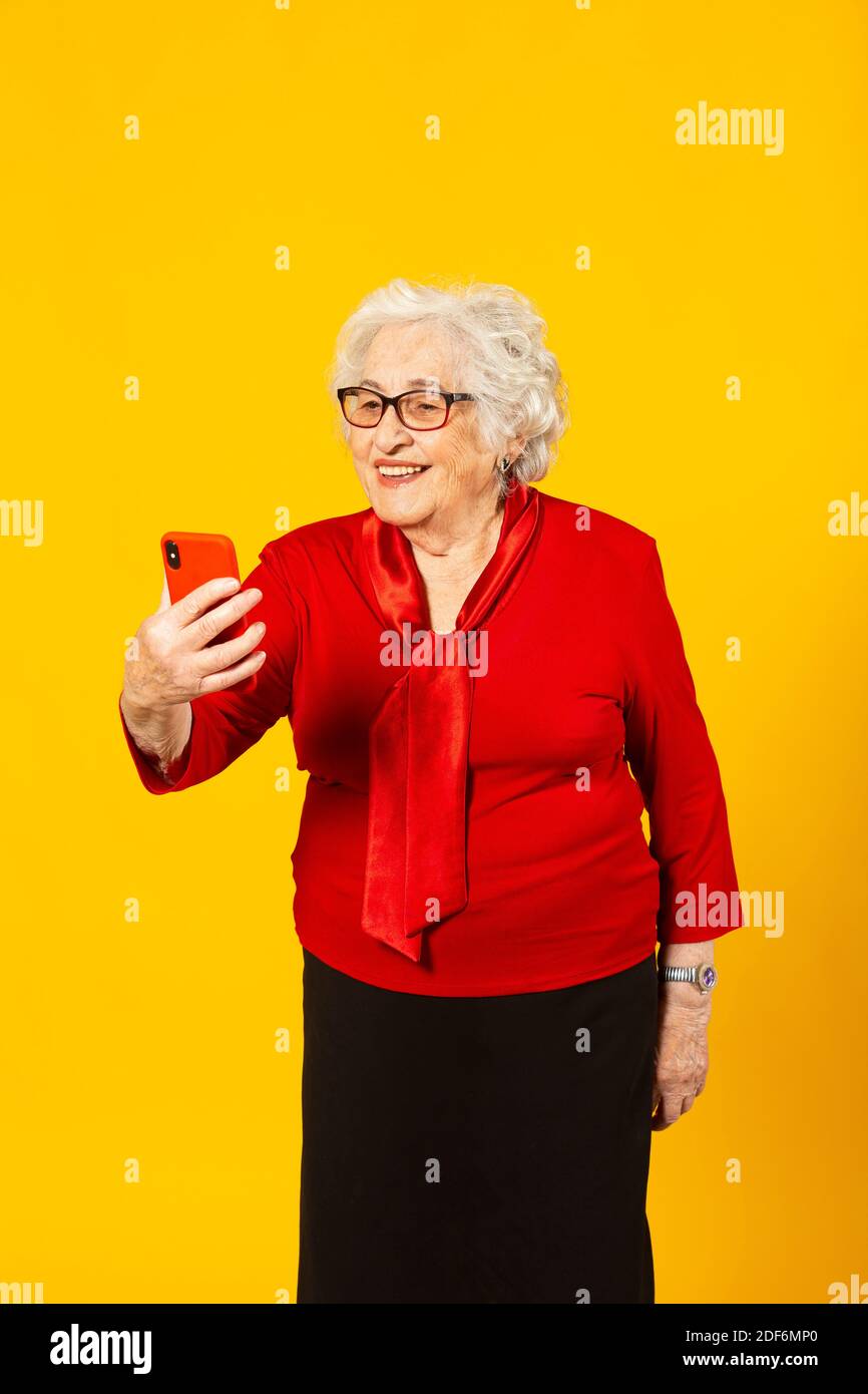 Portrait studio d'une femme âgée portant une chemise rouge sur fond jaune et passer un appel vidéo avec un téléphone mobile rouge Banque D'Images Portrait studio d'une femme âgée portant une chemise rouge sur fond jaune et passer un appel vidéo avec un téléphone mobile rouge Banque D'Images