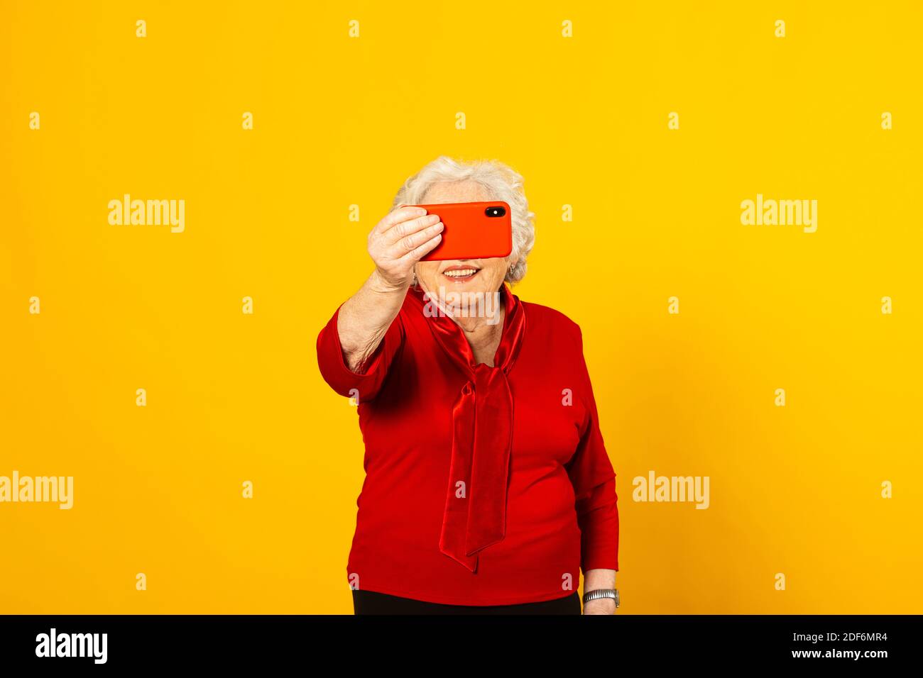 Portrait studio d'une femme âgée portant une chemise rouge sur fond jaune et passer un appel vidéo avec un téléphone mobile rouge Banque D'Images Portrait studio d'une femme âgée portant une chemise rouge sur fond jaune et passer un appel vidéo avec un téléphone mobile rouge Banque D'Images