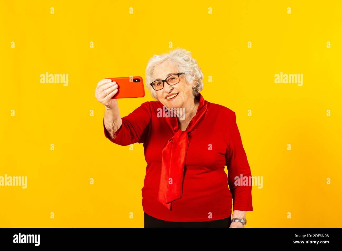Portrait studio d'une femme âgée portant une chemise rouge sur fond jaune et passer un appel vidéo avec un téléphone mobile rouge Banque D'Images Portrait studio d'une femme âgée portant une chemise rouge sur fond jaune et passer un appel vidéo avec un téléphone mobile rouge Banque D'Images