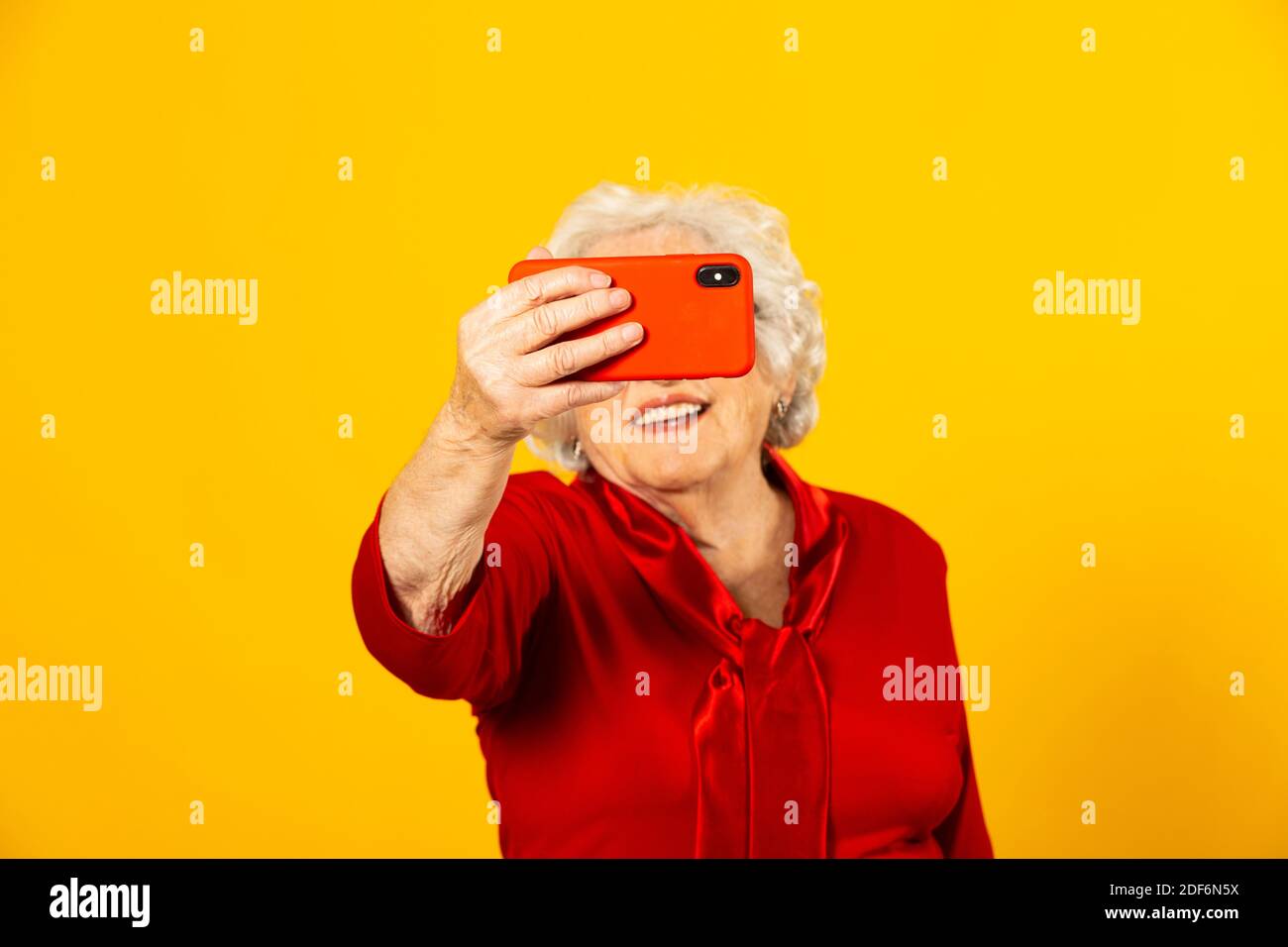 Portrait studio d'une femme âgée portant une chemise rouge sur fond jaune et passer un appel vidéo avec un téléphone mobile rouge Banque D'Images Portrait studio d'une femme âgée portant une chemise rouge sur fond jaune et passer un appel vidéo avec un téléphone mobile rouge Banque D'Images