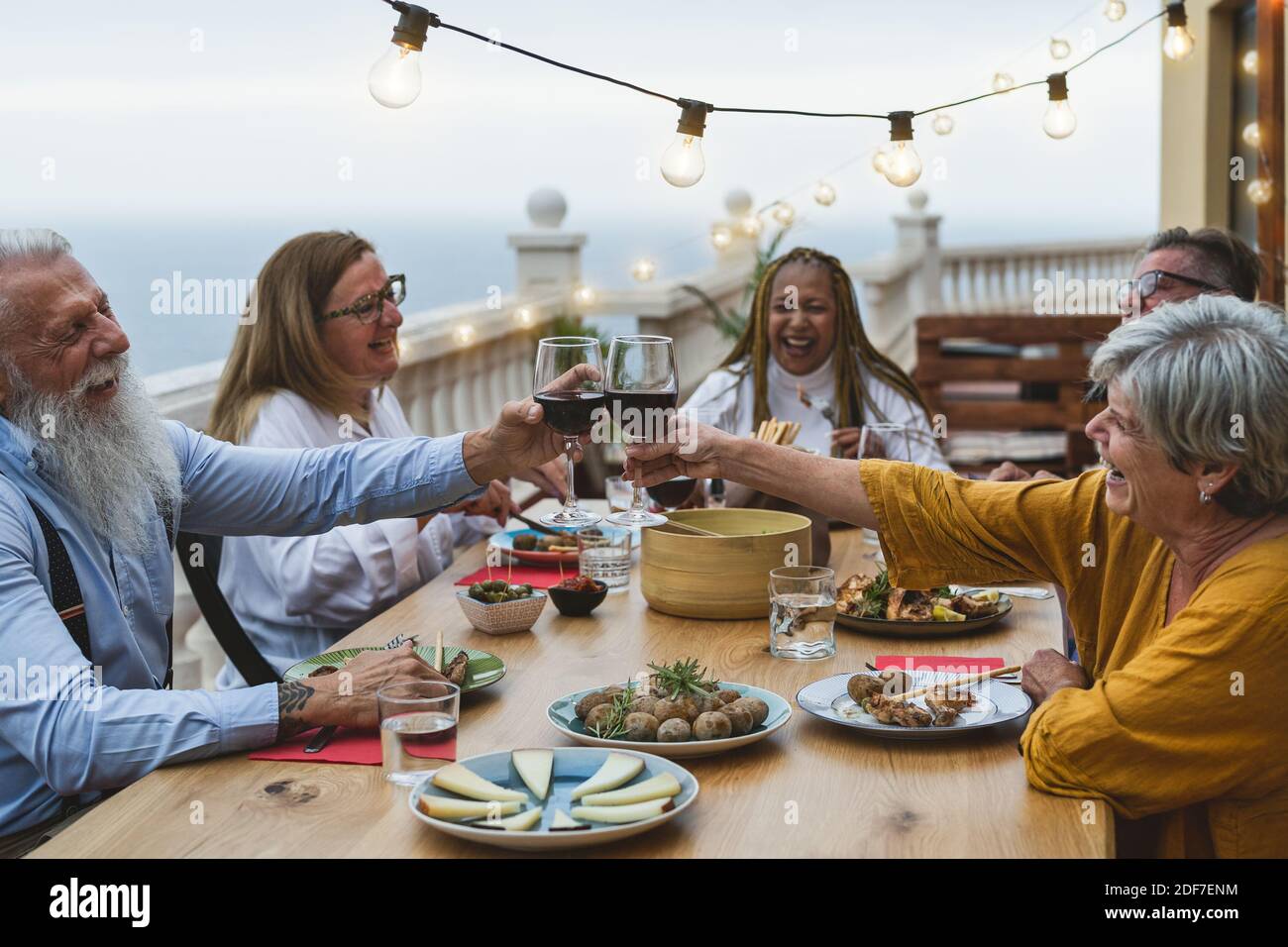 Des amis seniors multiraciaux s'amusent à manger ensemble et à faire du toaster avec Dîner avec vin rouge sur le patio de la maison - repas et vacances concept Banque D'Images