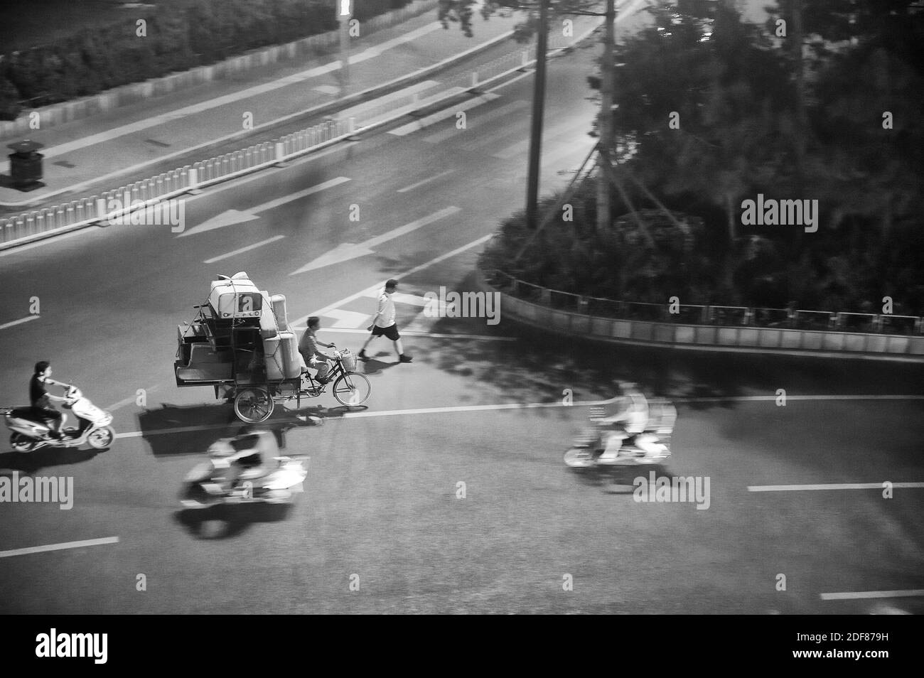 Man riding tricycle transportant une montagne de vieux meubles et du personnel. Au milieu d'un Carrefour en Chine. Photo en noir et blanc. Banque D'Images