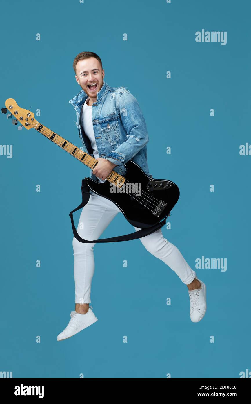 attrayant joueur de guitare fou homme en jean t-shirt avec guitare saut haut isolé sur fond bleu Banque D'Images