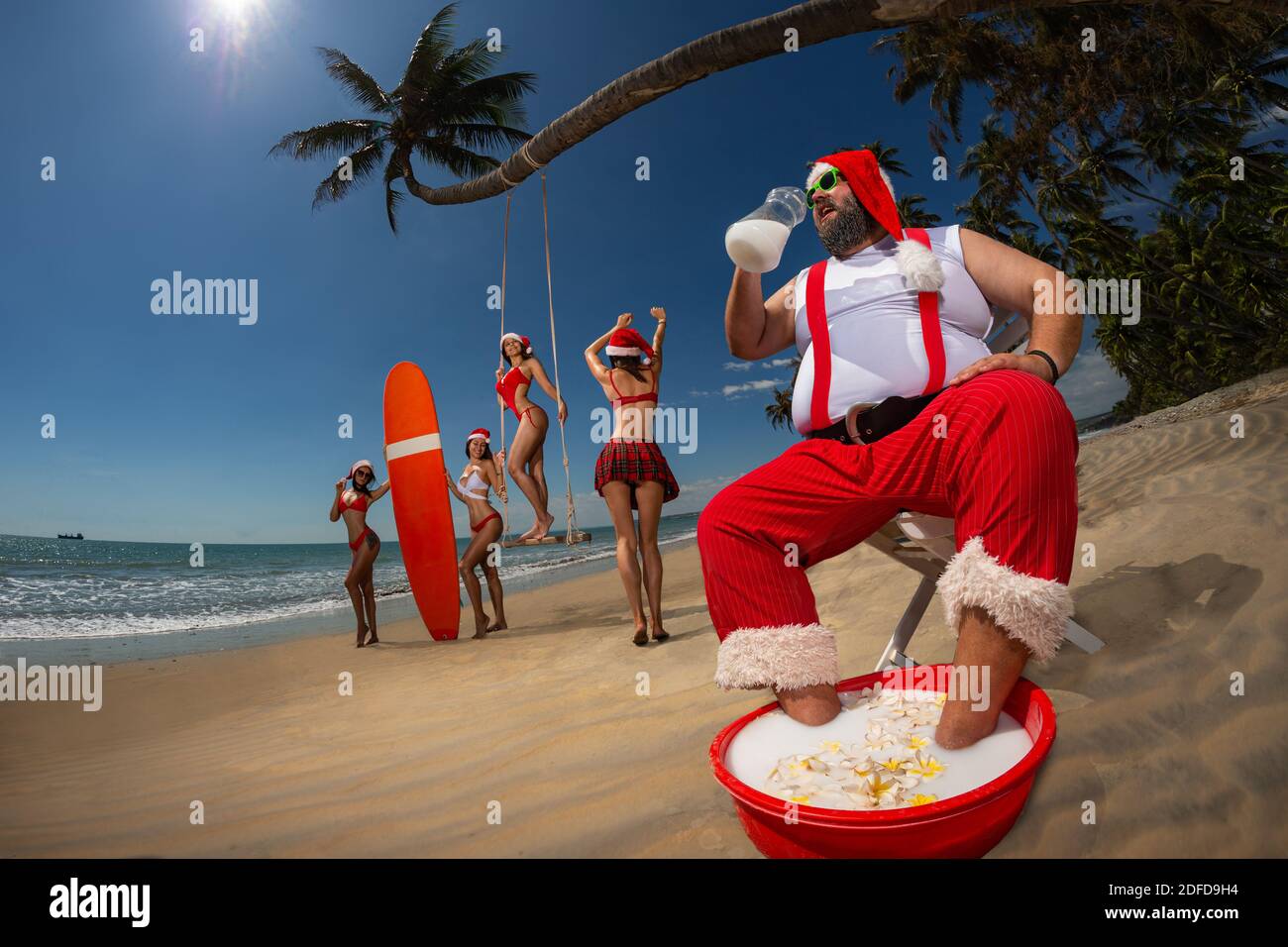 Noël Santa Claus a un repos sur un transat sur une plage tropicale de sable et de boire du lait de coco. De belles femmes s'amusent sur la plage. Banque D'Images