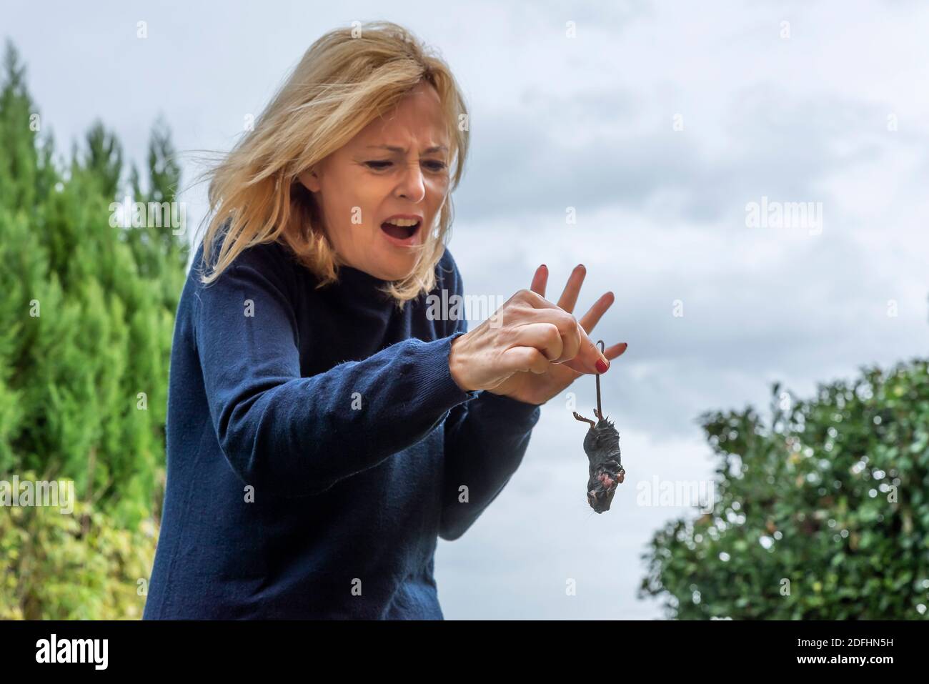 Une femme blonde d'âge moyen tient un rat humide près de la queue avec une expression terrifiée et dégoûtée Banque D'Images