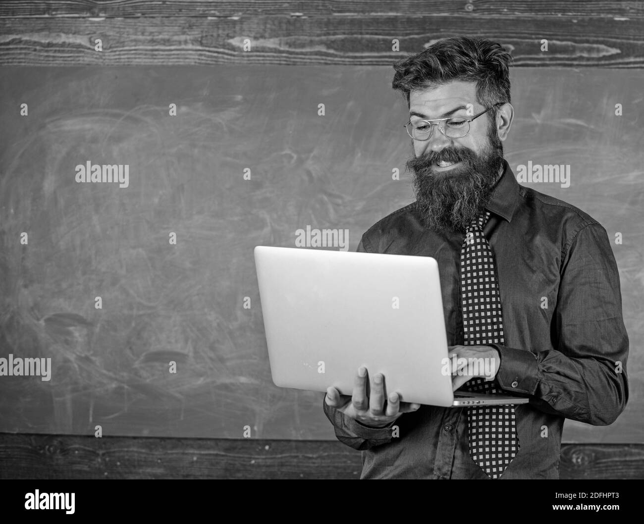 Surfer sur Internet. Homme barbu enseignant avec ordinateur portable moderne surf toile de fond de tableau noir. Hipster professeur porter lunettes et cravate tient ordinateur portable surfer sur Internet. Concept d'enseignement à distance. Banque D'Images