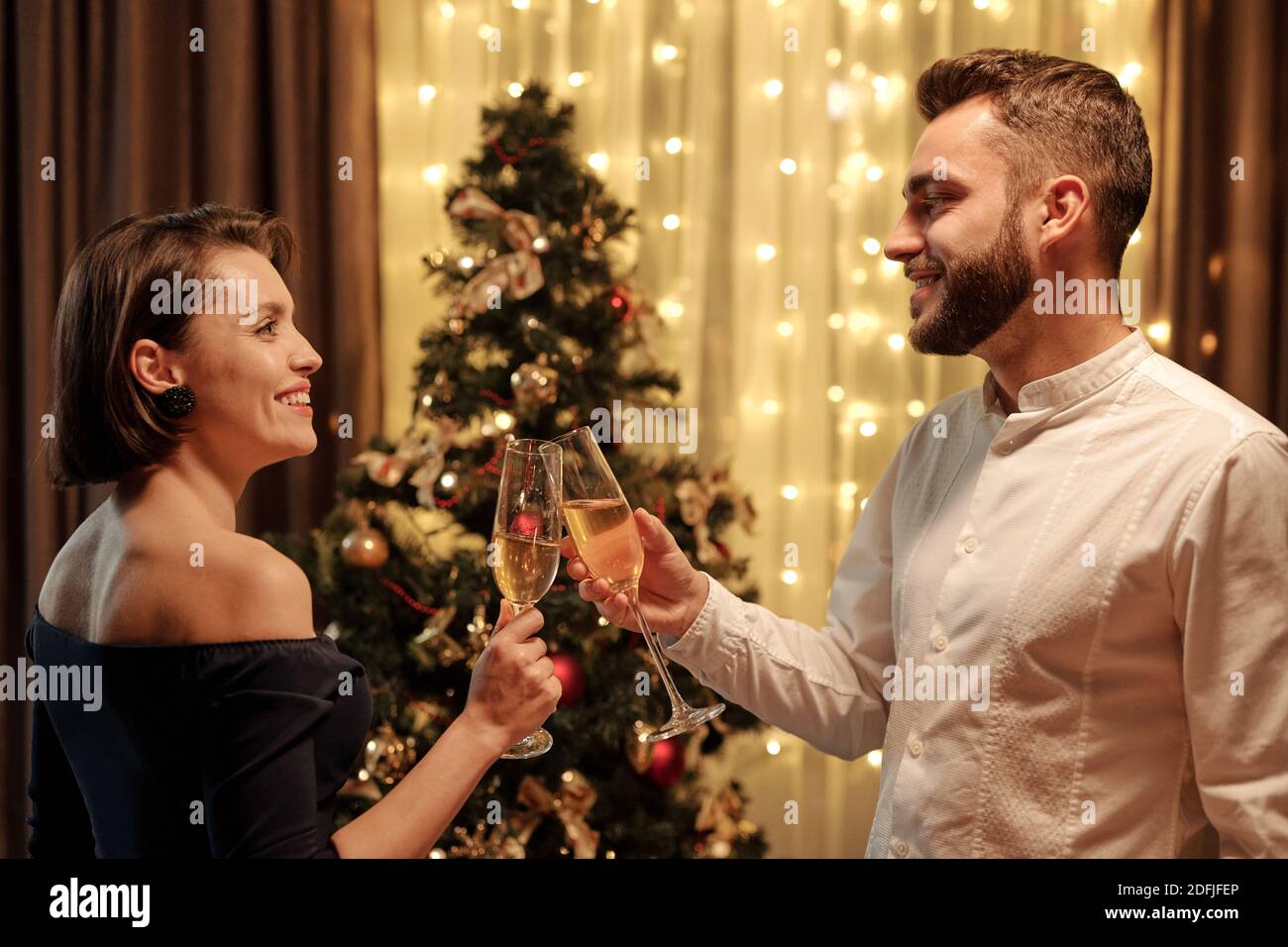Vue latérale d'un jeune couple joyeux se regardant l'un l'autre avec des sourires tout en se clinquant avec des flûtes de champagne devant de caméra par arbre de noël Banque D'Images