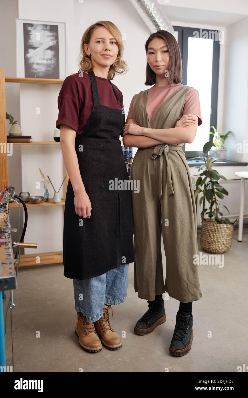 Deux jeunes artisans interculturels en vêtements de travail debout à côté de les uns les autres devant la caméra dans un grand atelier et je vous regarde Banque D'Images