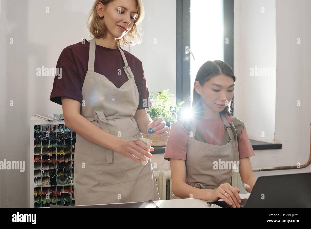 Jeune femme en tablier mettant la perle de verre faite à la main en petit sac en lin ou en coton pendant que sa collègue asiatique saisit des données dans un ordinateur portable Banque D'Images