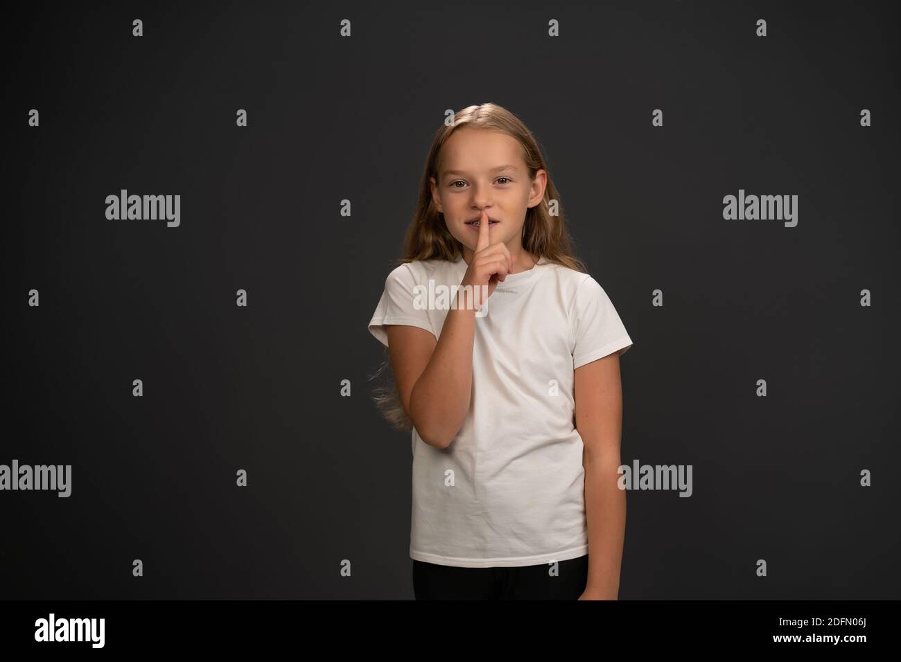 Une petite fille tient son doigt sur ses lèvres pour dire à tout le monde de garder le silence. Enfant portant un t-shirt blanc souriant à l'appareil photo isolé sur le gris foncé ou Banque D'Images