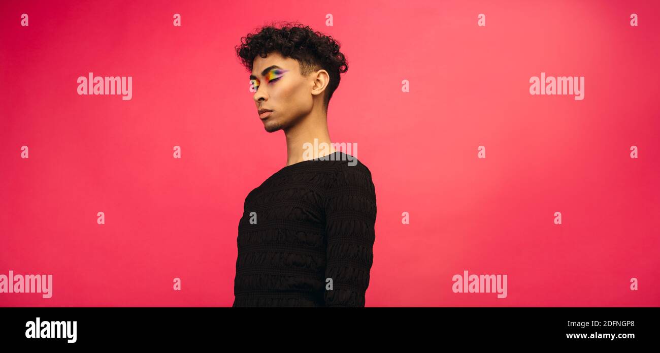 Jeune homme à l'ombre des yeux arc-en-ciel maquillage debout sur fond rouge avec ses yeux fermés. Homme fluide de sexe en t-shirt noir. Banque D'Images