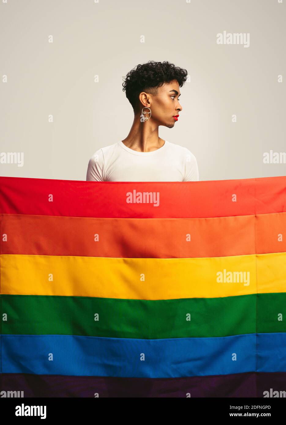 Jeune homme avec maquillage et boucles d'oreilles debout derrière le drapeau de fierté gay. Homme transgenre derrière le drapeau lgbtq. Banque D'Images