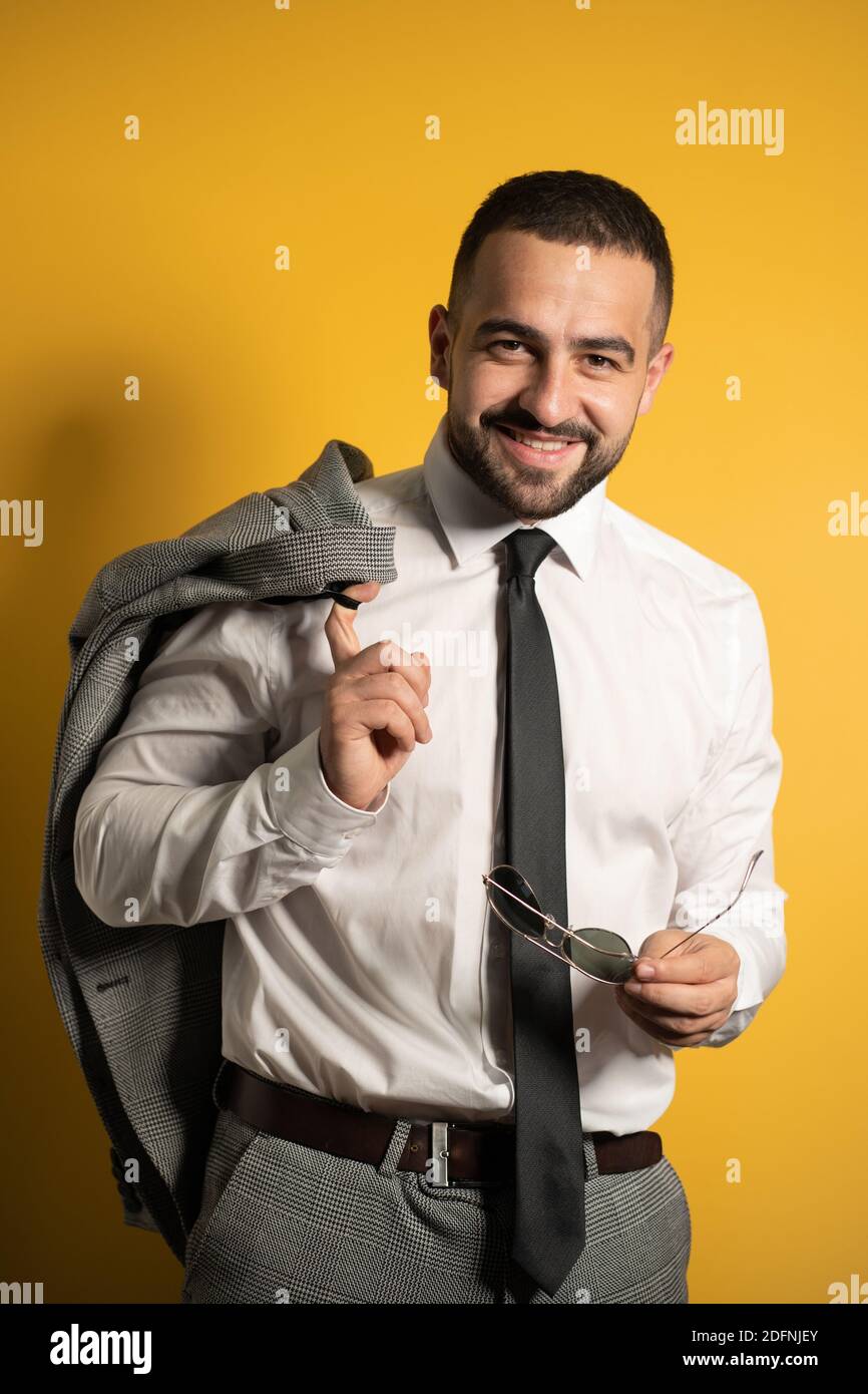 Beau jeune homme d'affaires tenant des lunettes de soleil dans la main et la veste sur son épaule isolée sur fond jaune Banque D'Images