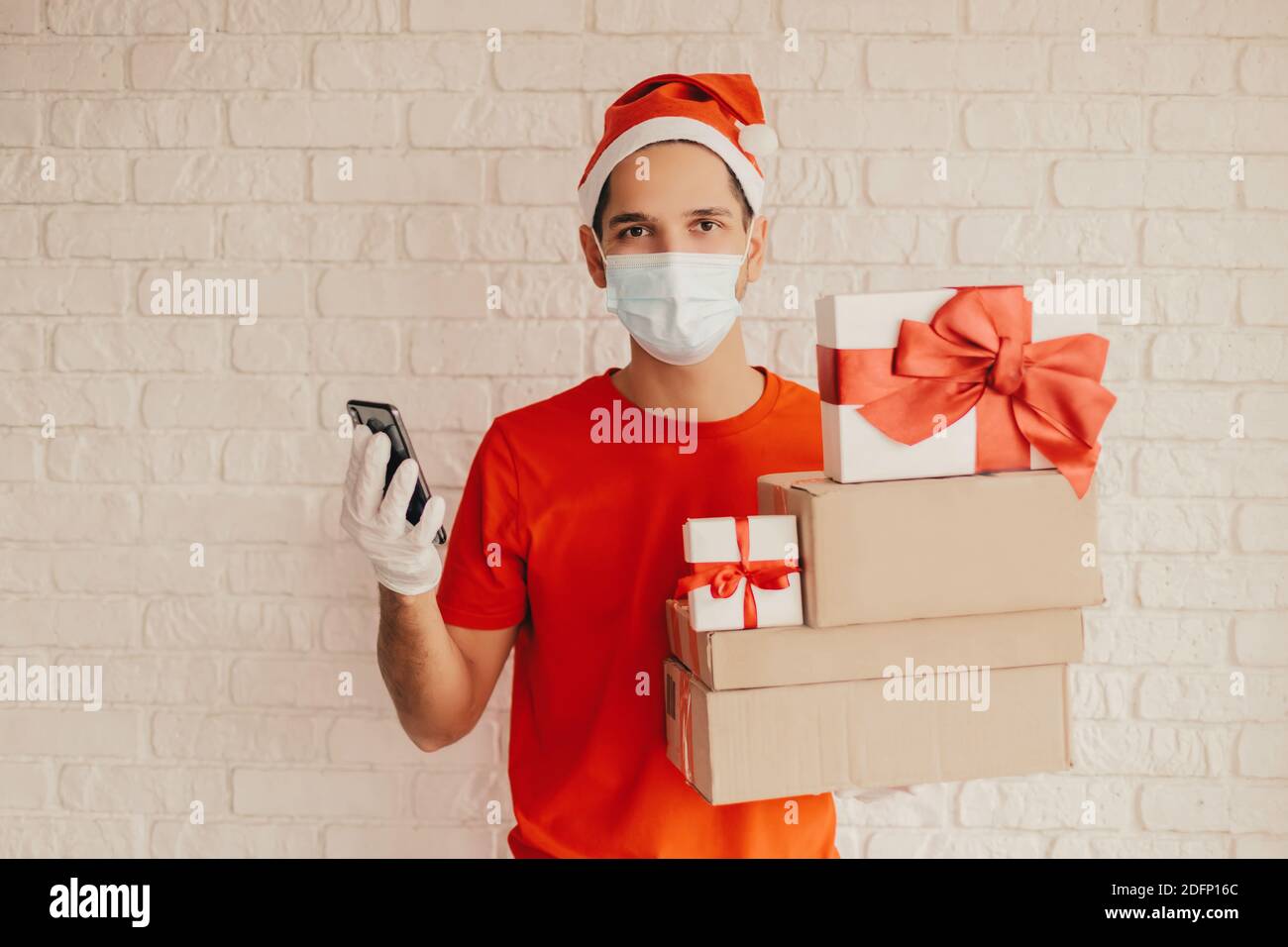 Jeune homme messagerie dans le masque médical, gants de protection, chapeau de père noël livraison des cadeaux de Noël. Bon liveur dans le masque tenir le smartphone, cadeau, Banque D'Images