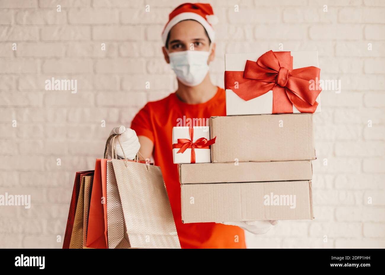 Bon liveur dans le masque médical, chapeau de père Noël, gant de protection porte sacs à provisions, boîtes-cadeaux en carton avec noeud rouge. Jeune homme messagerie en face Banque D'Images