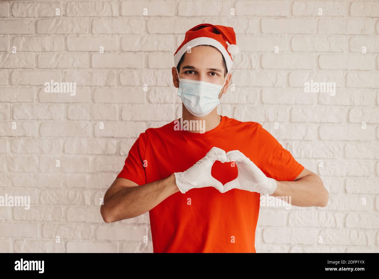 Jeune homme messagerie dans un masque médical, gants de protection, symbole d'amour de geste de santa Hat avec les doigts, sourire. Bon liveur dans le masque du visage tenir hea Banque D'Images