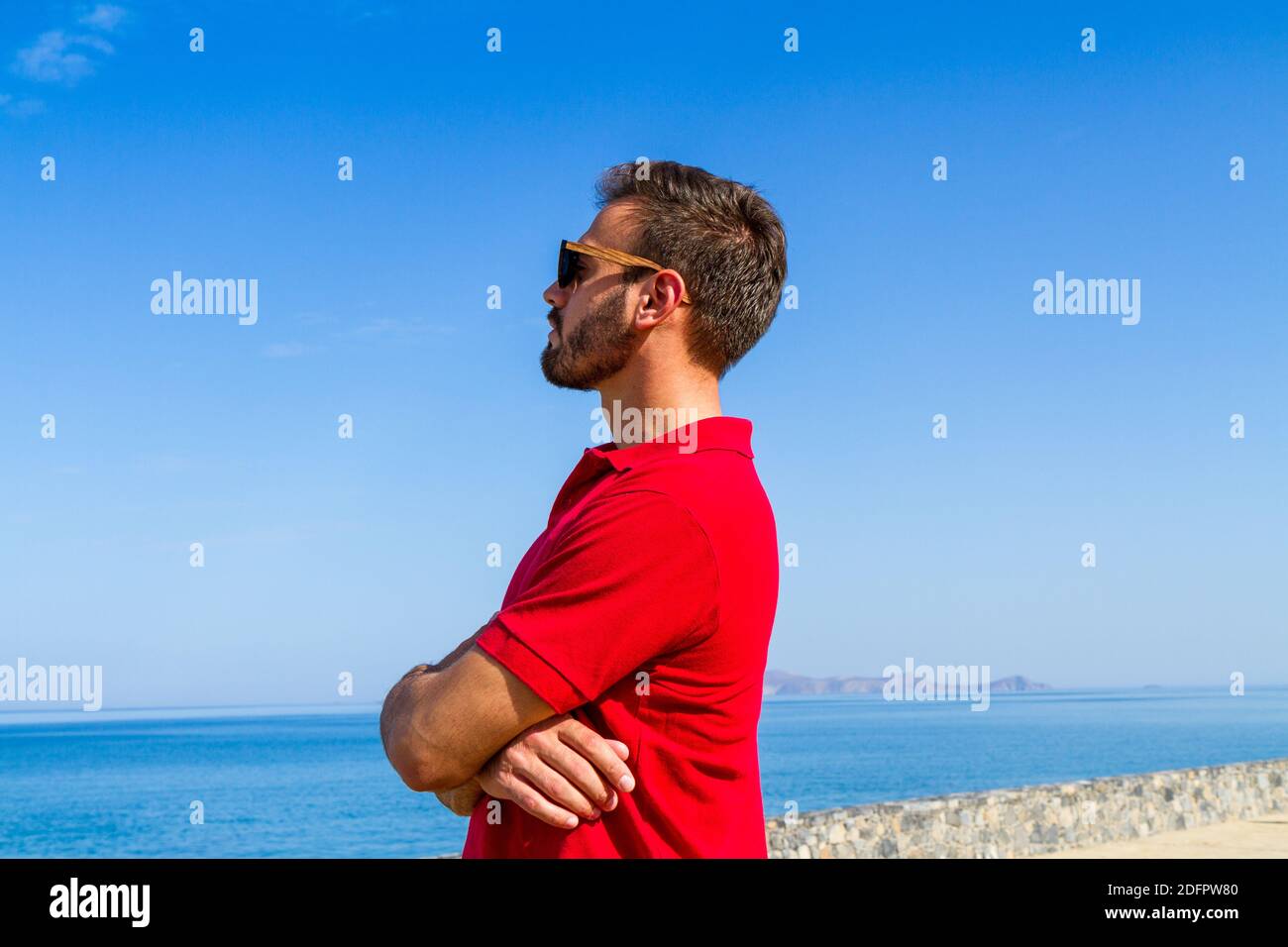 Jeune homme expressif dynamique ambitieux réussi barbu en t-shirt rouge lunettes de soleil en été bleu ciel océan fond. Concept de liberté de style de vie Banque D'Images