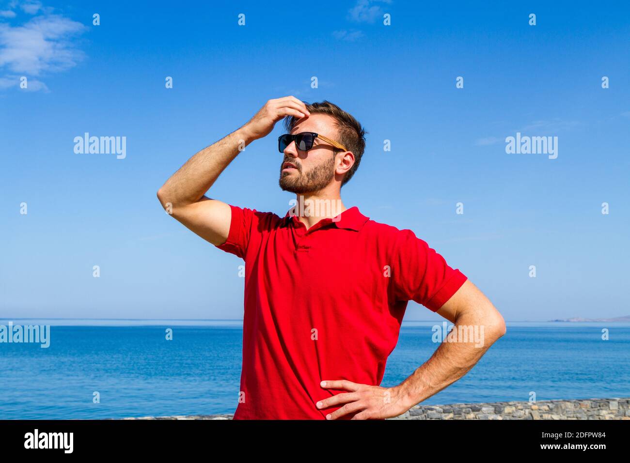 Jeune homme expressif dynamique ambitieux réussi barbu en t-shirt rouge lunettes de soleil en été bleu ciel océan fond. Concept de liberté de style de vie Banque D'Images