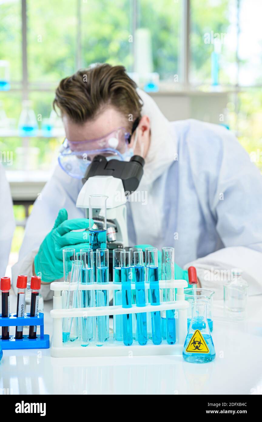 Équipement de laboratoire avec liquide bleu à l'intérieur du support sur la table Avec un scientifique mâle portant une combinaison de protection travaillant avec le microscope pour vaccin de recherche à l Banque D'Images