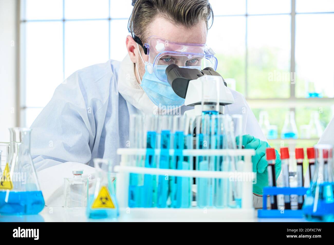 Un scientifique mâle portant une combinaison de protection travaillant avec le microscope et de nombreux équipements de laboratoire pour la recherche de vaccins en laboratoire. Chercheur médical spécialisé Banque D'Images