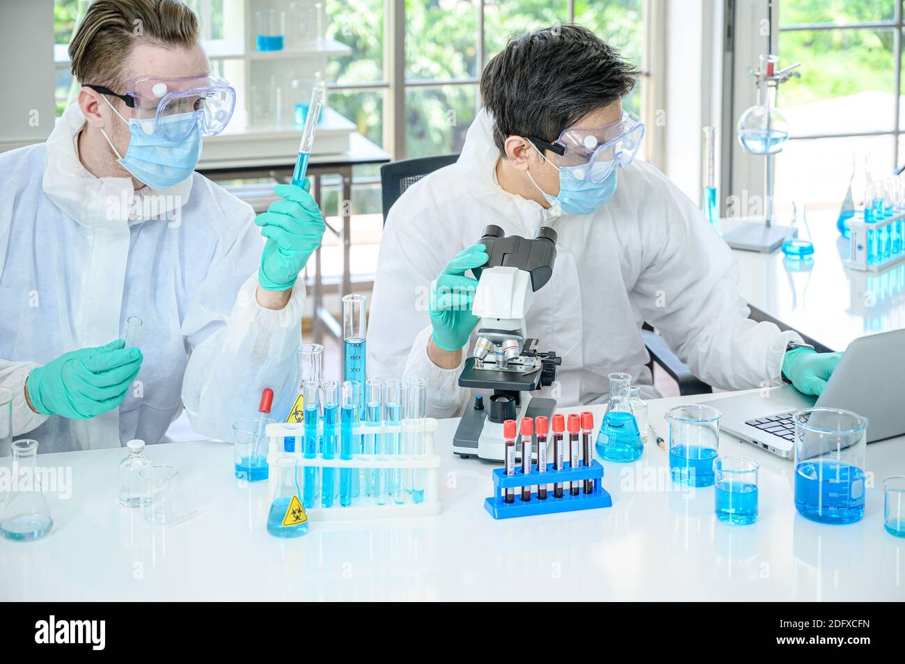 Couple de scientifiques mâles portant une combinaison de protection travaillant avec le microscope et Ordinateur portable et de nombreux équipements de laboratoire pour la recherche du vaccin contre le coronavirus à Banque D'Images