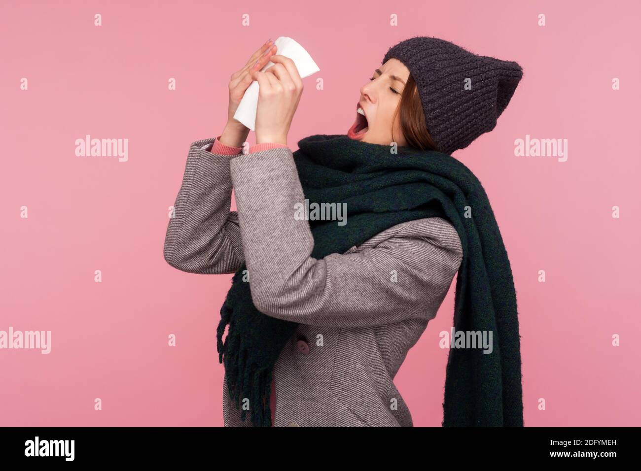 Contrarié frustrée femme dans le chapeau et le foulard largement ouvert sa bouche allant éternuer dans la serviette à ses mains, souffrant du froid et de la grippe. Studio intérieur sho Banque D'Images