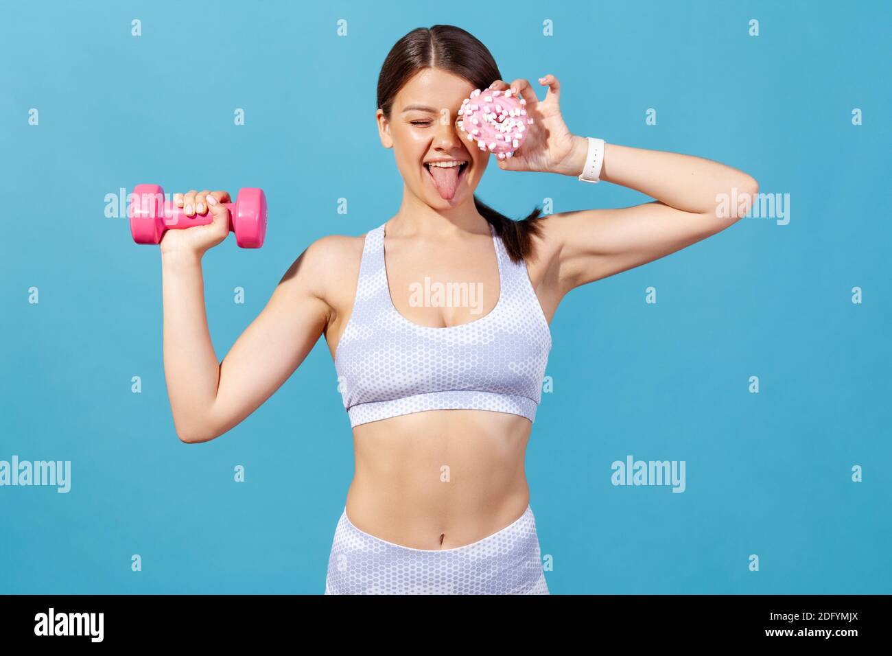 Drôle de sport positif femme dans les collants et le haut avoir l'amusement, la fermeture de l'œil avec le donut et montrant la langue, tenant la haltère dans la main, brûlant des calories supplémentaires. Banque D'Images