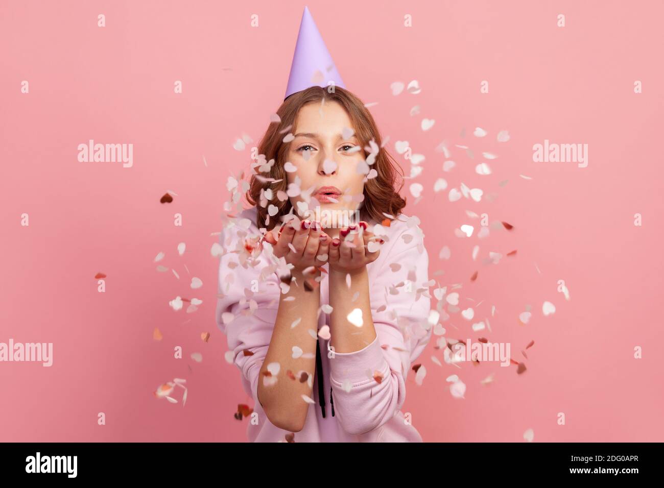 Portrait d'une adolescente enjouée insouciante en sweat à capuche et en forme de cône de fête soufflant en forme de coeur confetti, appréciant anniversaire ou jour de la Saint Valentin, ambiance festive. Banque D'Images