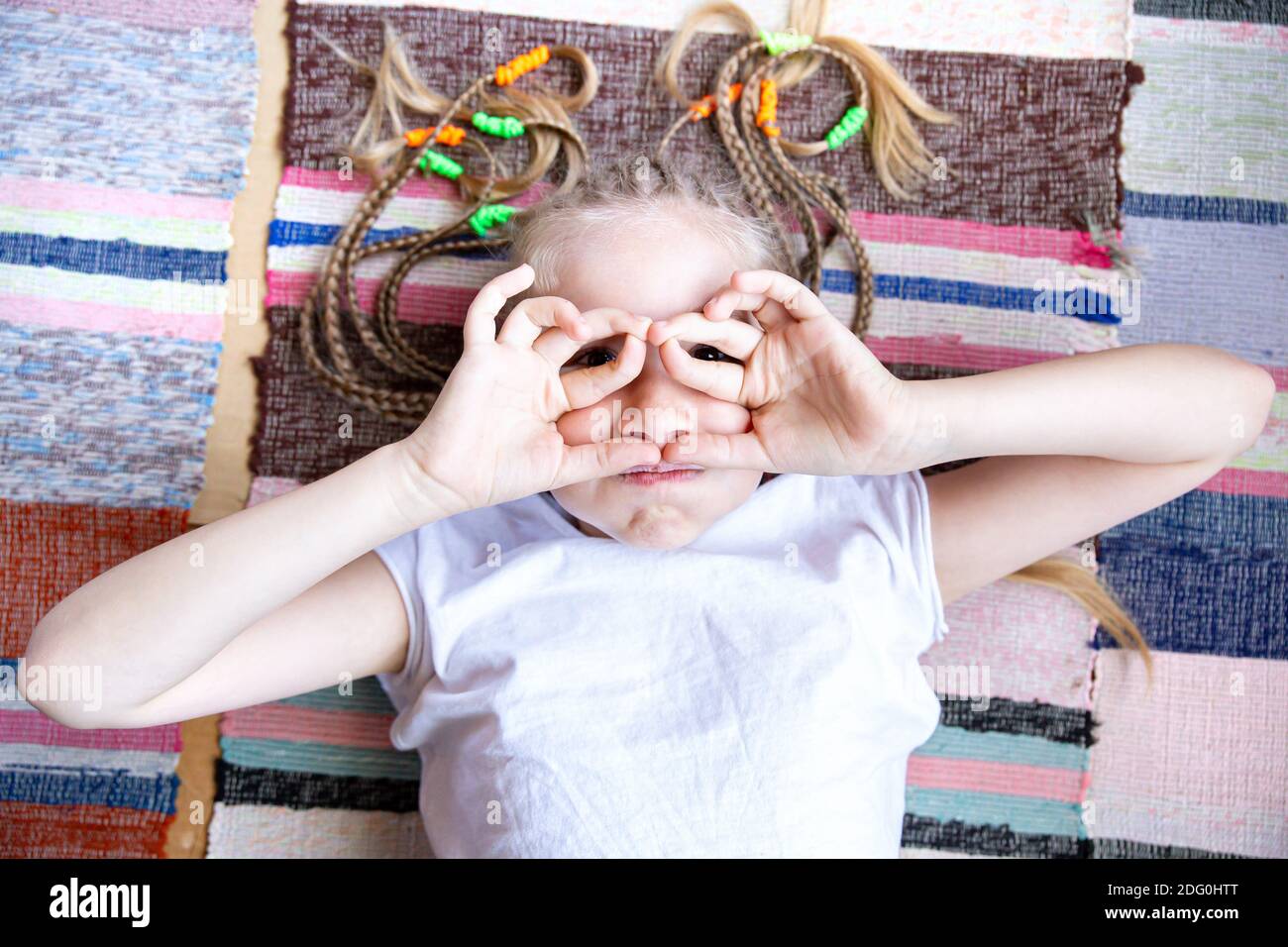 Portrait en gros plan une jeune fille gaie avec des queues de porc, repose sur un tapis tissé rayé, sourires, lunettes des doigts pliés. Banque D'Images
