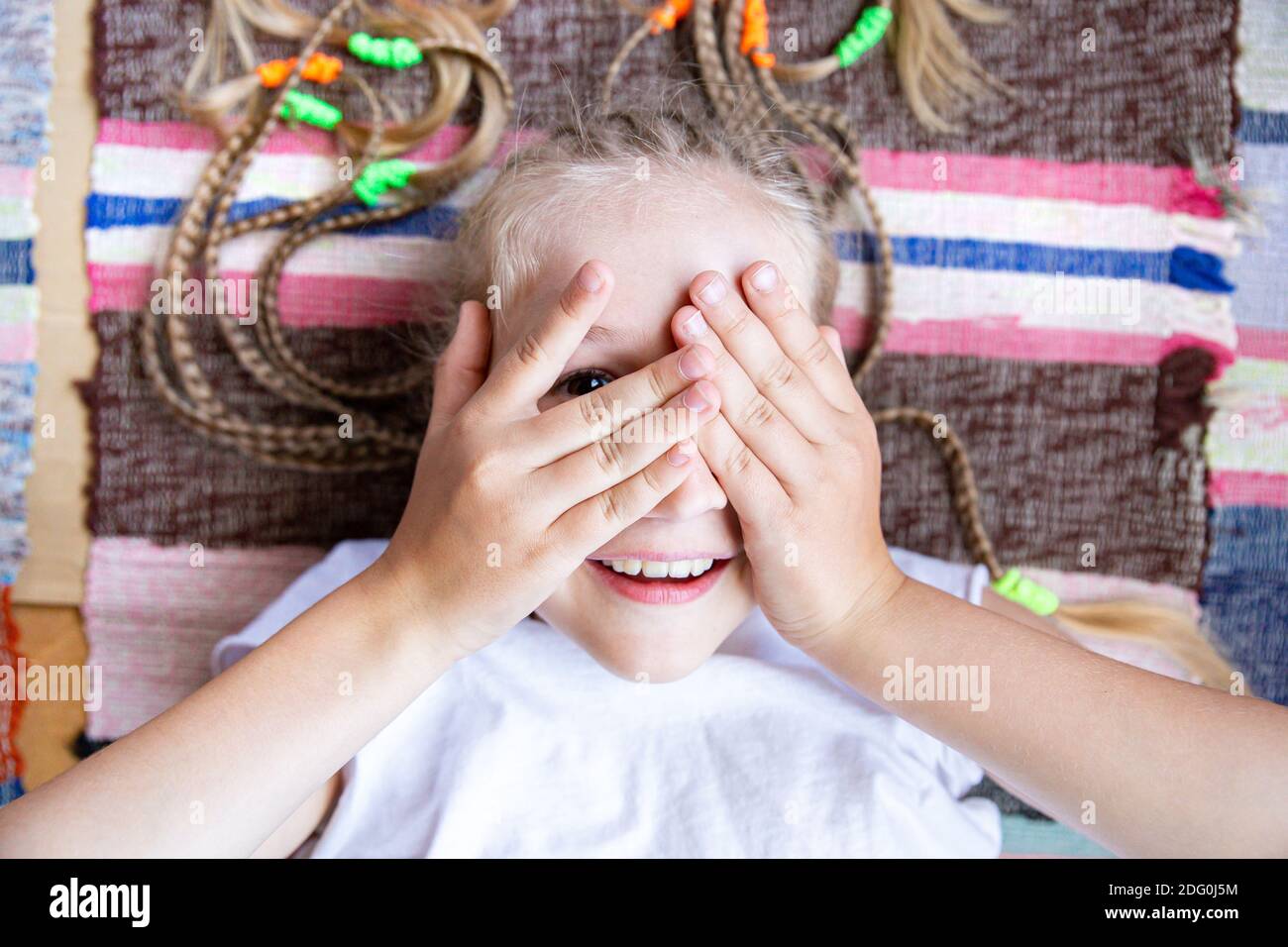 Gros plan portrait d'une jeune fille gaie avec des queues de porc couchées sur un tapis tissé rayé, souriant, jouant cacher et chercher. Banque D'Images