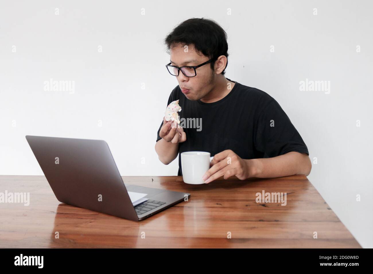 Un jeune homme asiatique mange du Donut et boit du café tout en travaillant avec un ordinateur portable. Indonésie Homme porter chemise noire fond gris isolé. Banque D'Images