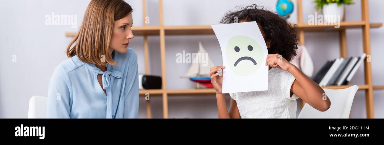 Fille afro-américaine autiste couvrant le visage avec une expression malheureuse sur papier près du psychologue sur fond flou, bannière Banque D'Images