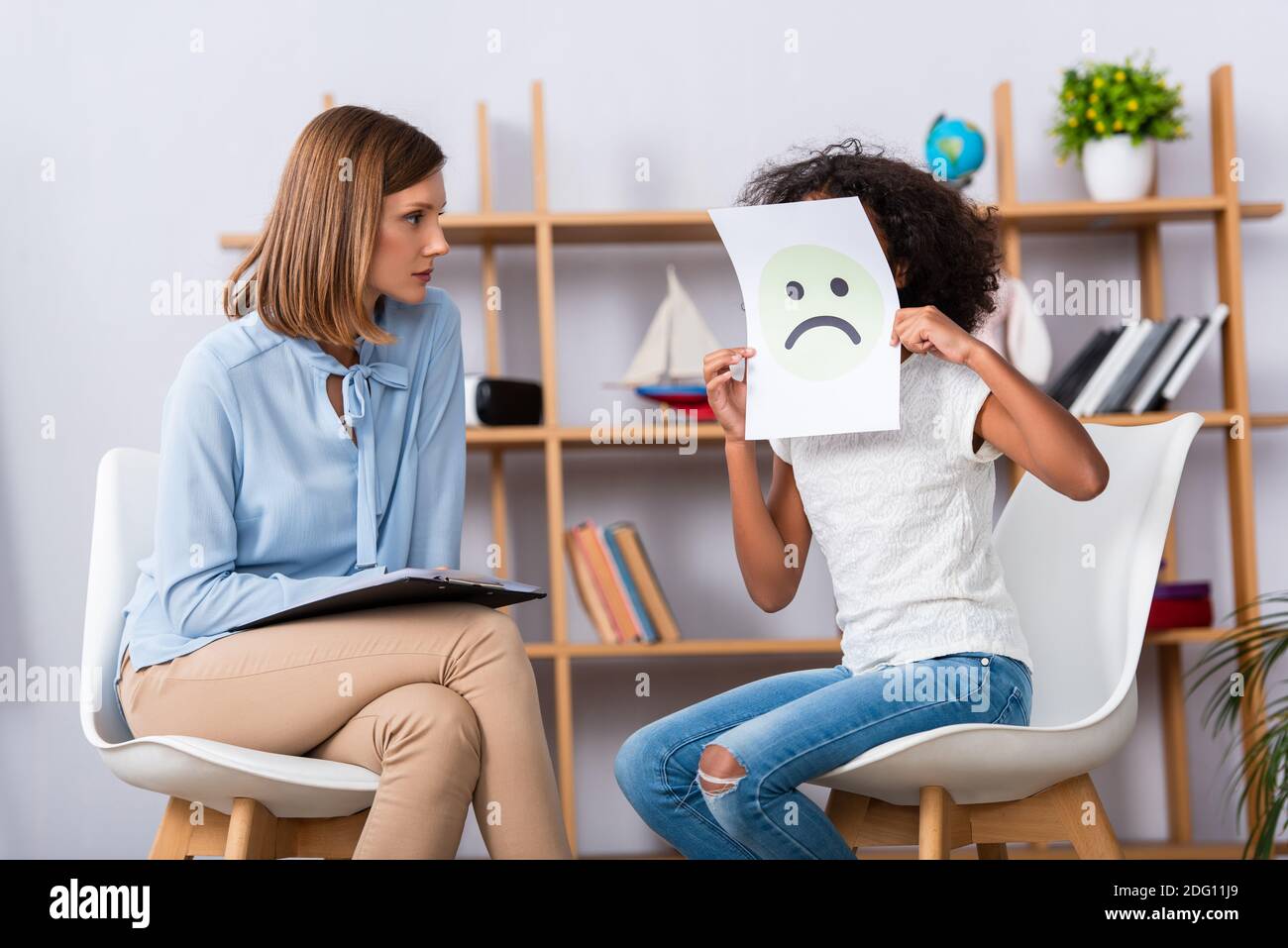 Psychologue regardant une fille afro-américaine couvrant le visage avec malheureux expression sur papier avec bureau flou en arrière-plan Banque D'Images