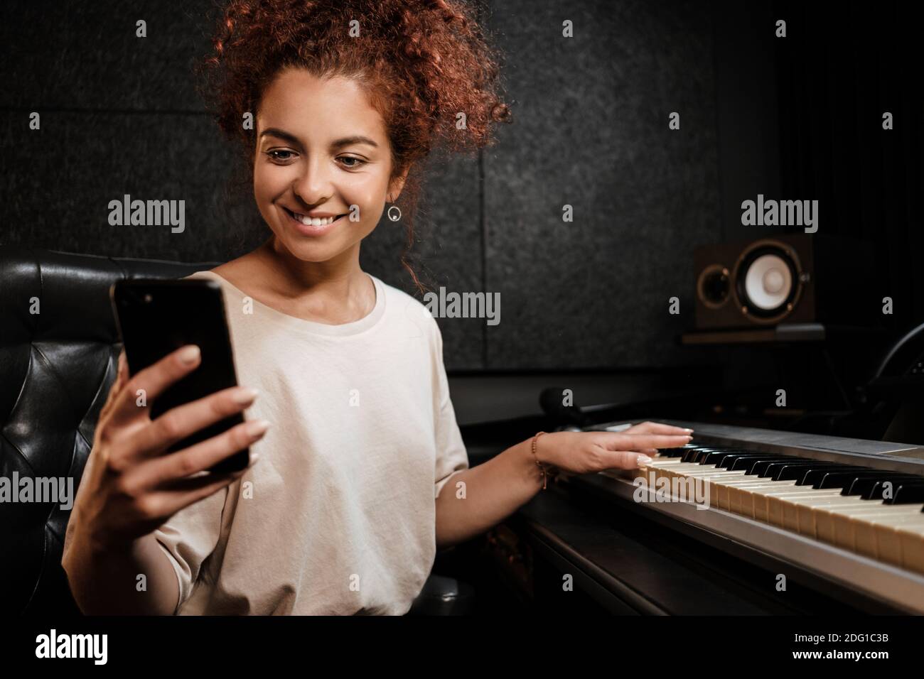 Jeune femme joyeuse utilisant le téléphone jouant sur le piano électrique en studio d'enregistrement Banque D'Images
