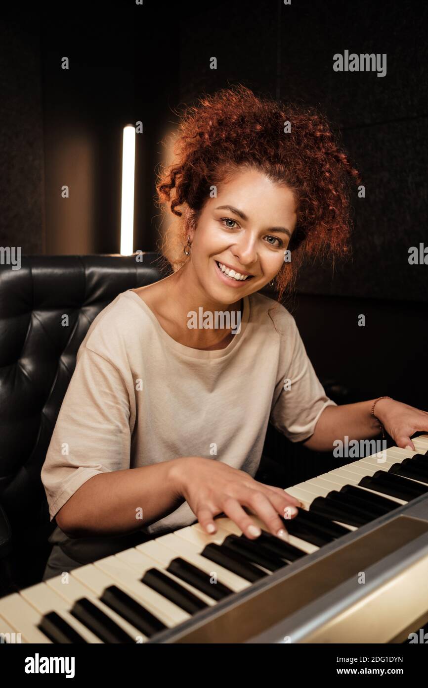 Belle fille joyeusement à la recherche dans l'appareil photo jouant sur piano électrique en studio. Musicien attrayant travaillant en studio d'enregistrement Banque D'Images