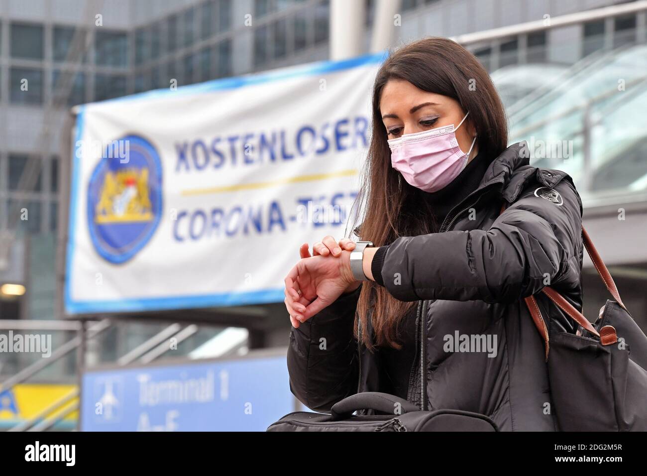 Voyages à thème en période de pandémie de corona. Jeune femme avec masque, masque à l'aéroport de Munich le 6 décembre 2020. Elle sort du terminal avec ses bagages et sa valise, derrière elle se trouve un panneau D'ESSAI GRATUIT CORONA. Elle regarde sa montre. MODÈLE DISPONIBLE ! | utilisation dans le monde entier Banque D'Images