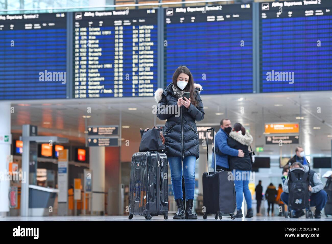 Voyages à thème en période de pandémie de corona. Jeune femme avec masque, masque avec sa valise, bagages à l'aéroport de Munich le 6 décembre 2020. Regarde son smartphone - derrière elle se trouve le tableau des départs. MODÈLE DISPONIBLE ! | utilisation dans le monde entier Banque D'Images
